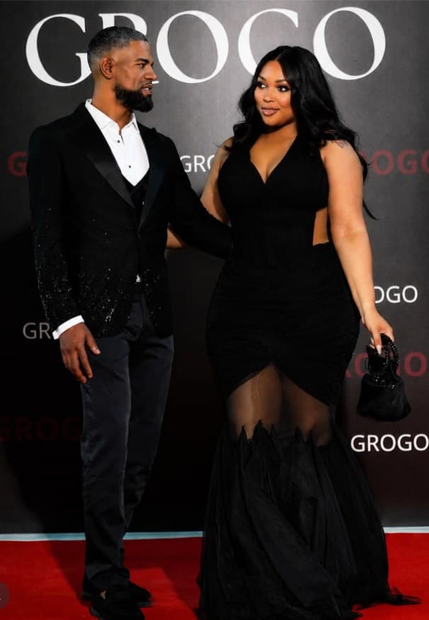 A couple on a red carpet; she wears a black gown with sheer panels