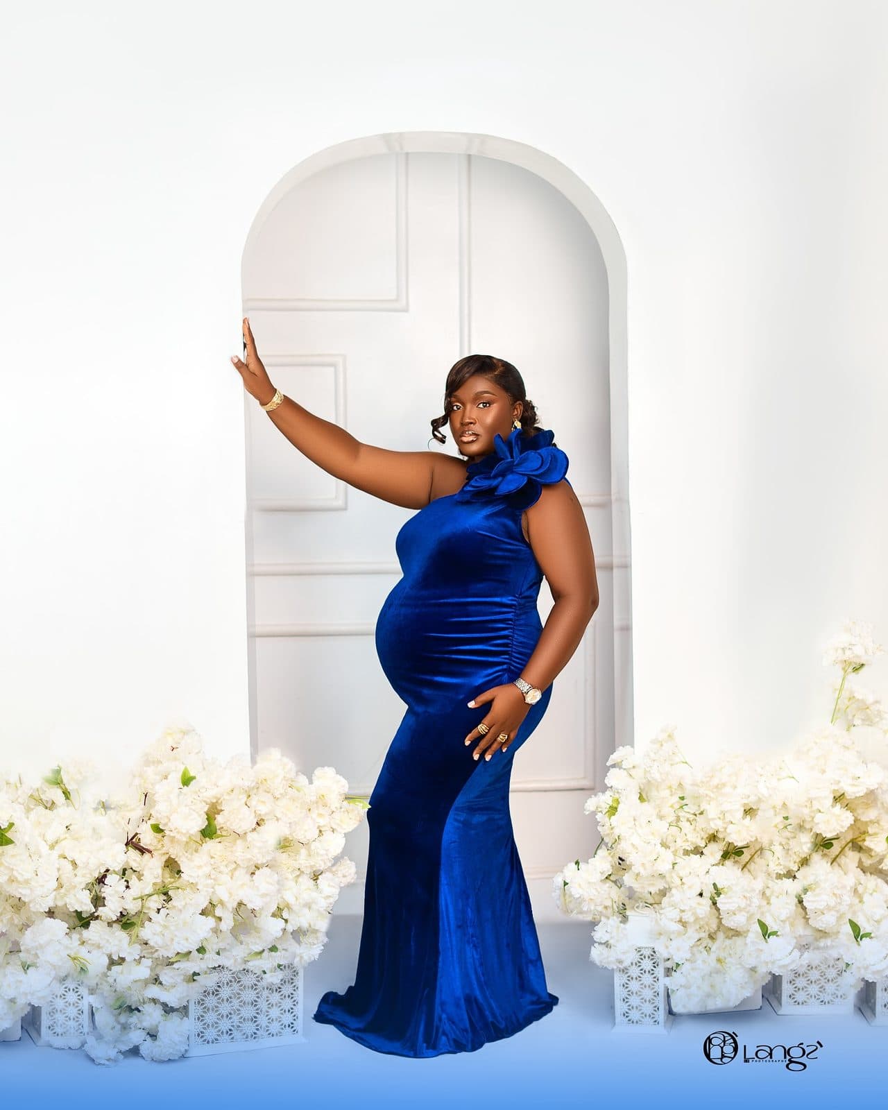 A woman in a royal-blue velvet maternity gown under a floral arch